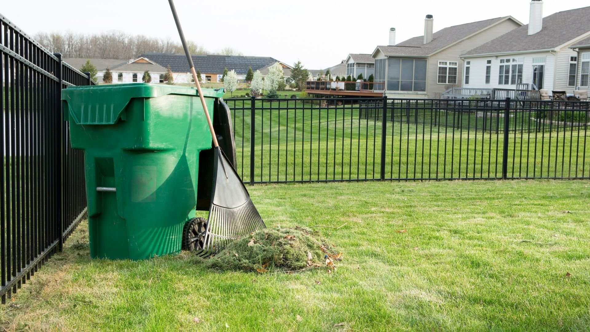 Moving green trash bin with rake and grass clippings in suburban backyard